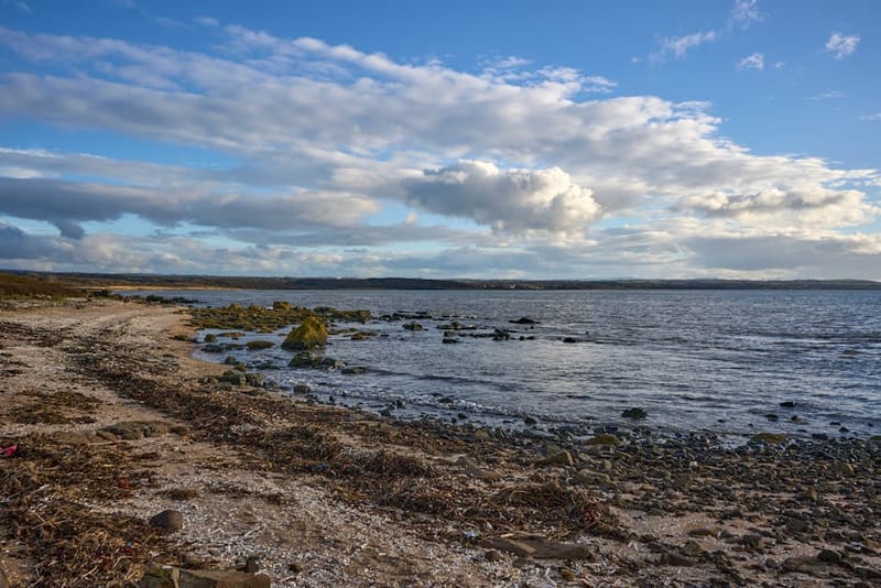 Serene view of rocky shoreline at Gosford Bay, Scotland under a blue sky and clouds.